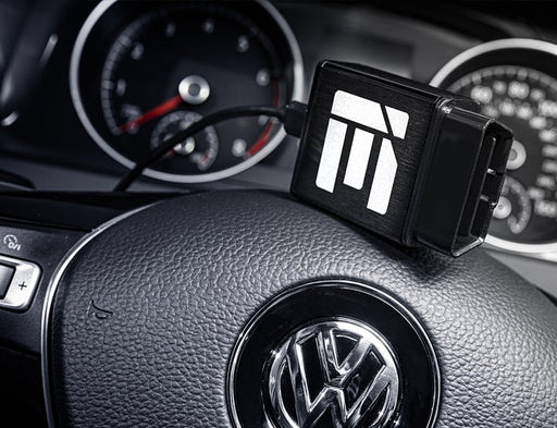 A close-up of a Volkswagen steering wheel featuring a black device with a white logo, plugged into the vehicle's diagnostic port.