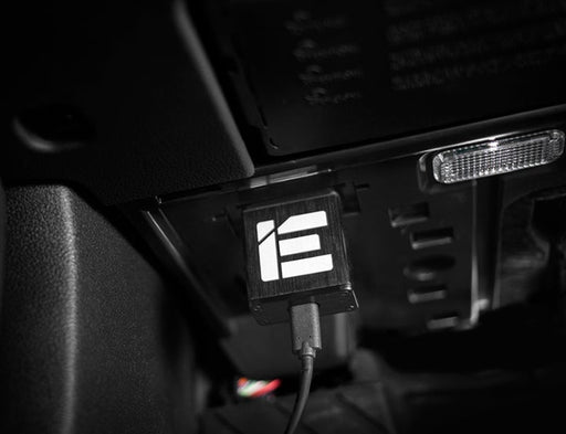 A close-up of a black electronic device with a white logo, plugged into a vehicle's dashboard, surrounded by dark interior elements.